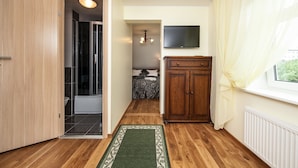 A hallway with a wooden door, a shower area, a bed with a patterned bedspread, a wooden cabinet, and a television mounted on the wall. - Embrace Guestrooms & Apartments (Parnu)