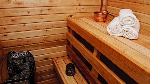 A wooden sauna room with benches, towels, a copper bucket, and a container of rocks. - Embrace Guestrooms & Apartments (Parnu)