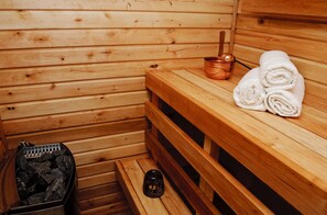 A wooden sauna room with benches, towels, a copper bucket, and a container of rocks.