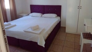 Een keurig opgemaakt bed met witte lakens en handdoeken, een paars hoofdeinde, een witte nachtkastje en een witte kledingkast in een hotelkamer.