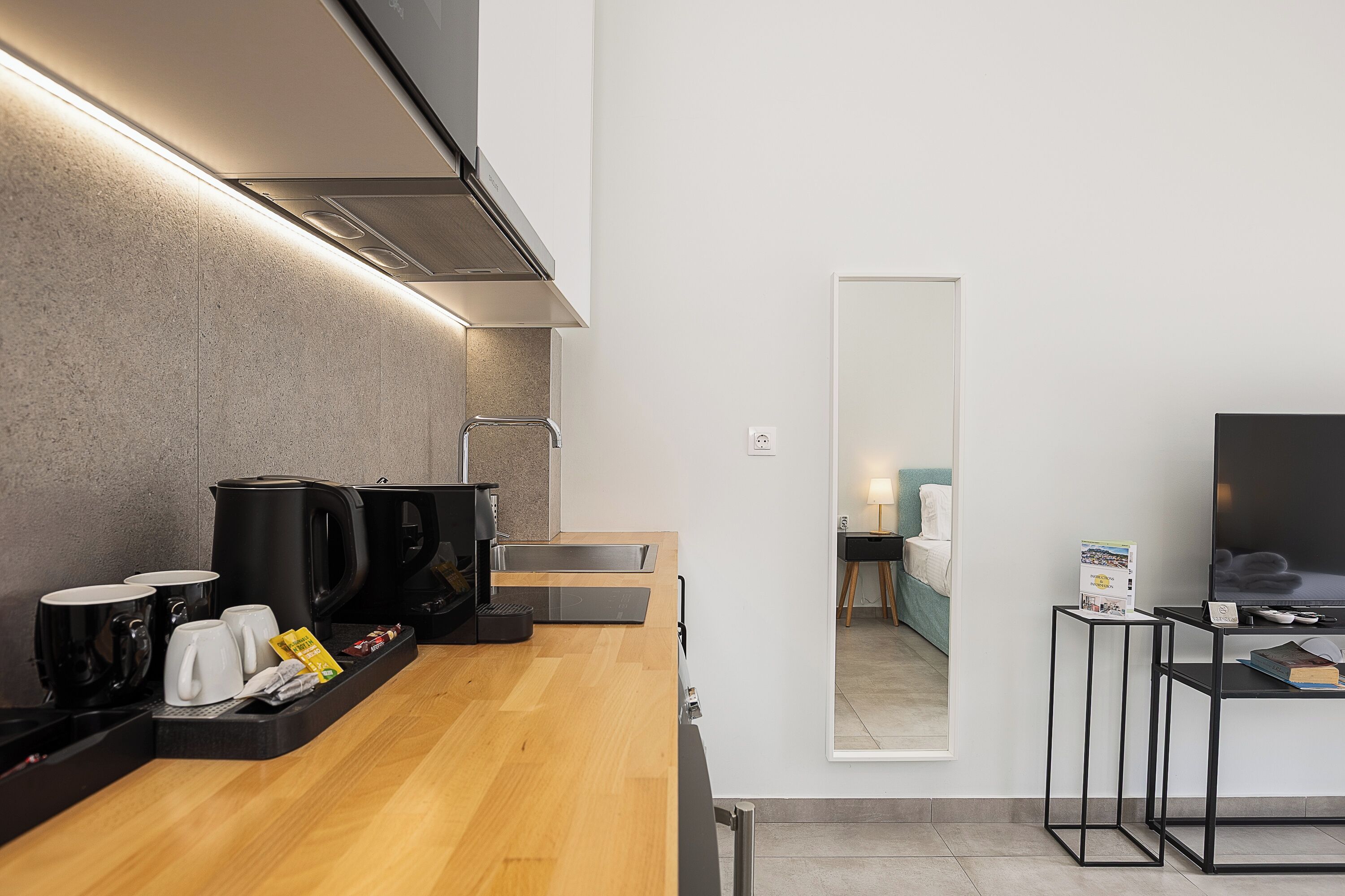A modern kitchen with a coffee station, a flat-screen TV, and a mirror reflecting a bedroom.