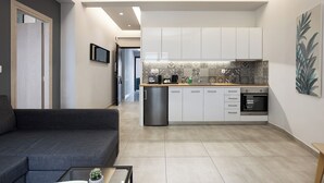 A modern kitchen with white cabinets, a built-in oven, and a refrigerator. There is a grey sofa and a coffee table with a plant in the living area. - The Omnia Collection Pagrati (Athens)