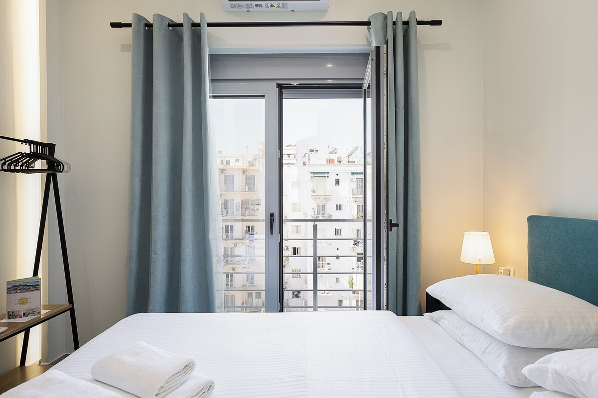 A hotel room with a bed, a nightstand with a lamp, a window with curtains, and a view of buildings outside.