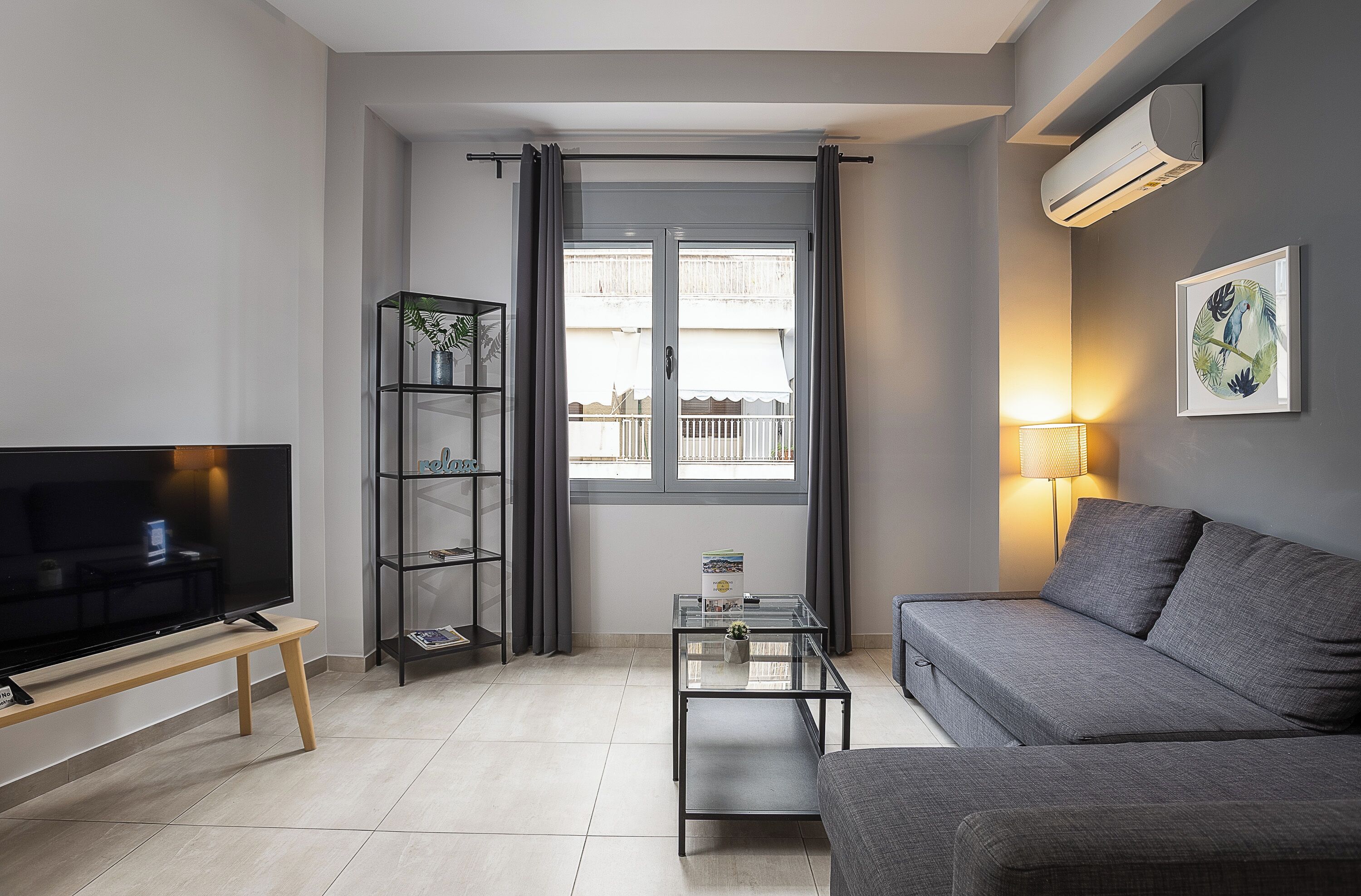 A modern living room with a grey sofa, a glass coffee table, a TV on a wooden stand, a bookshelf, and a window with curtains.