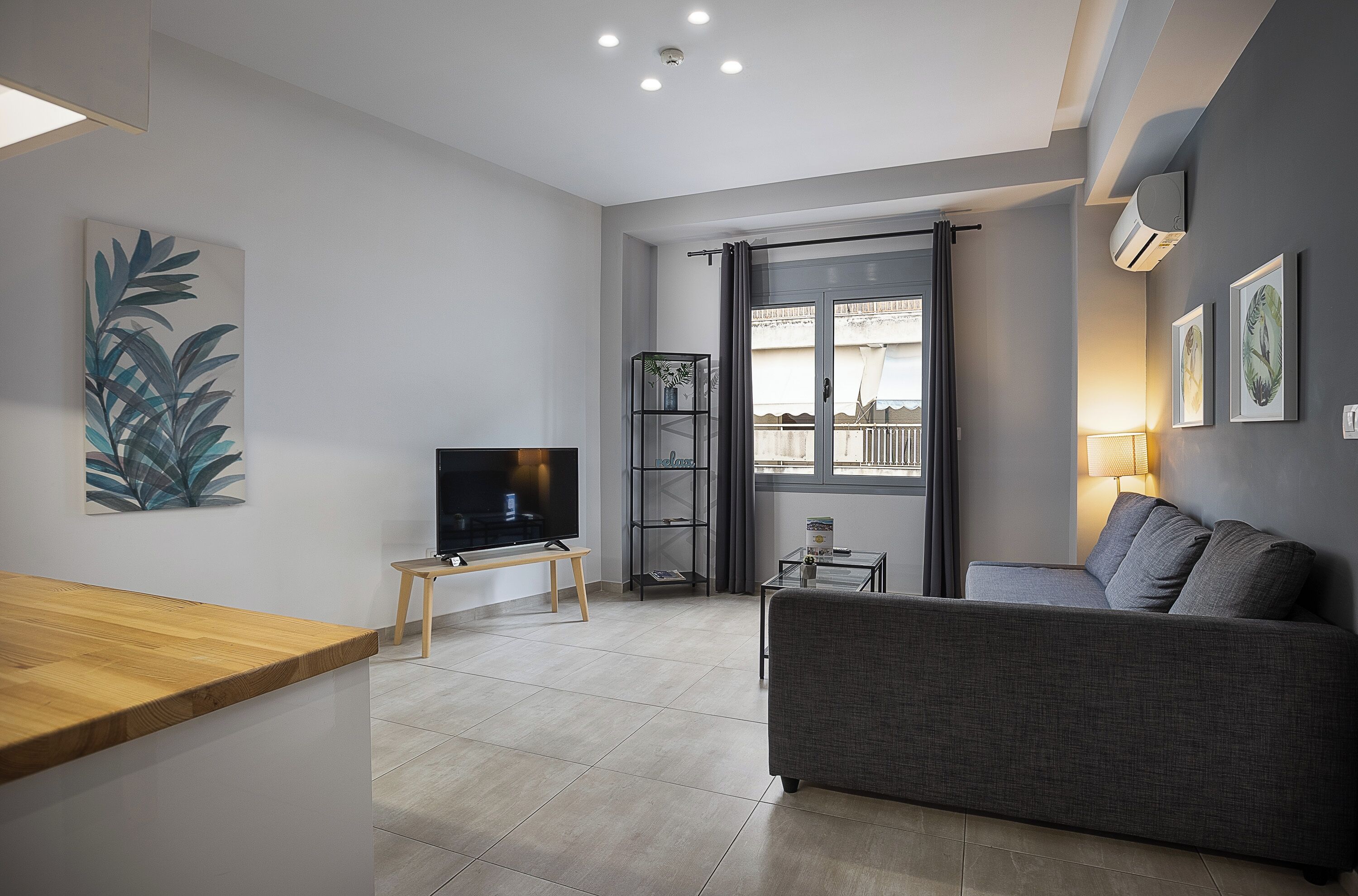 A modern living room with a grey sofa, a wooden coffee table, a TV on a stand, and a bookshelf.