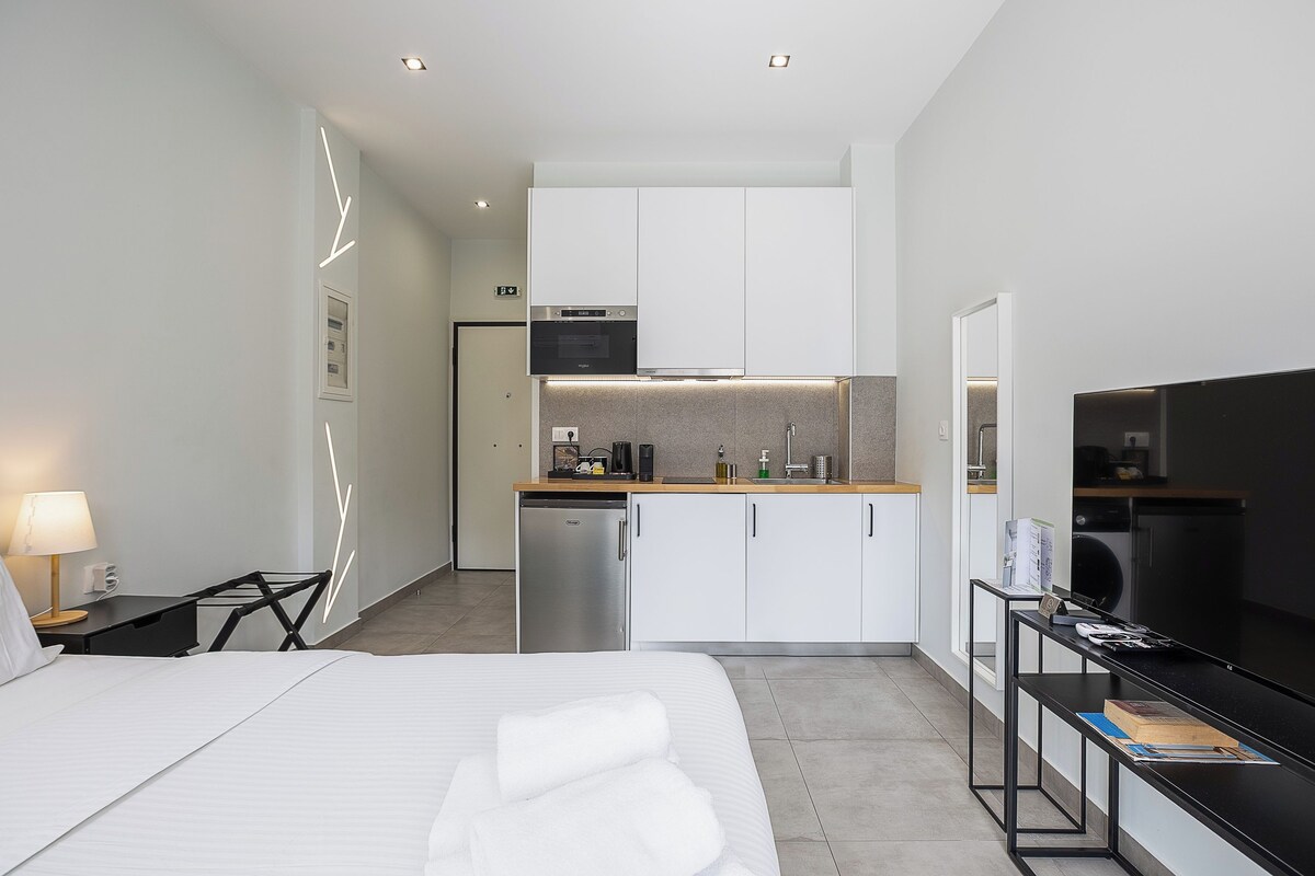 A modern hotel room with a bed, bedside table, lamp, and a kitchenette area including a microwave, oven, and coffee maker.