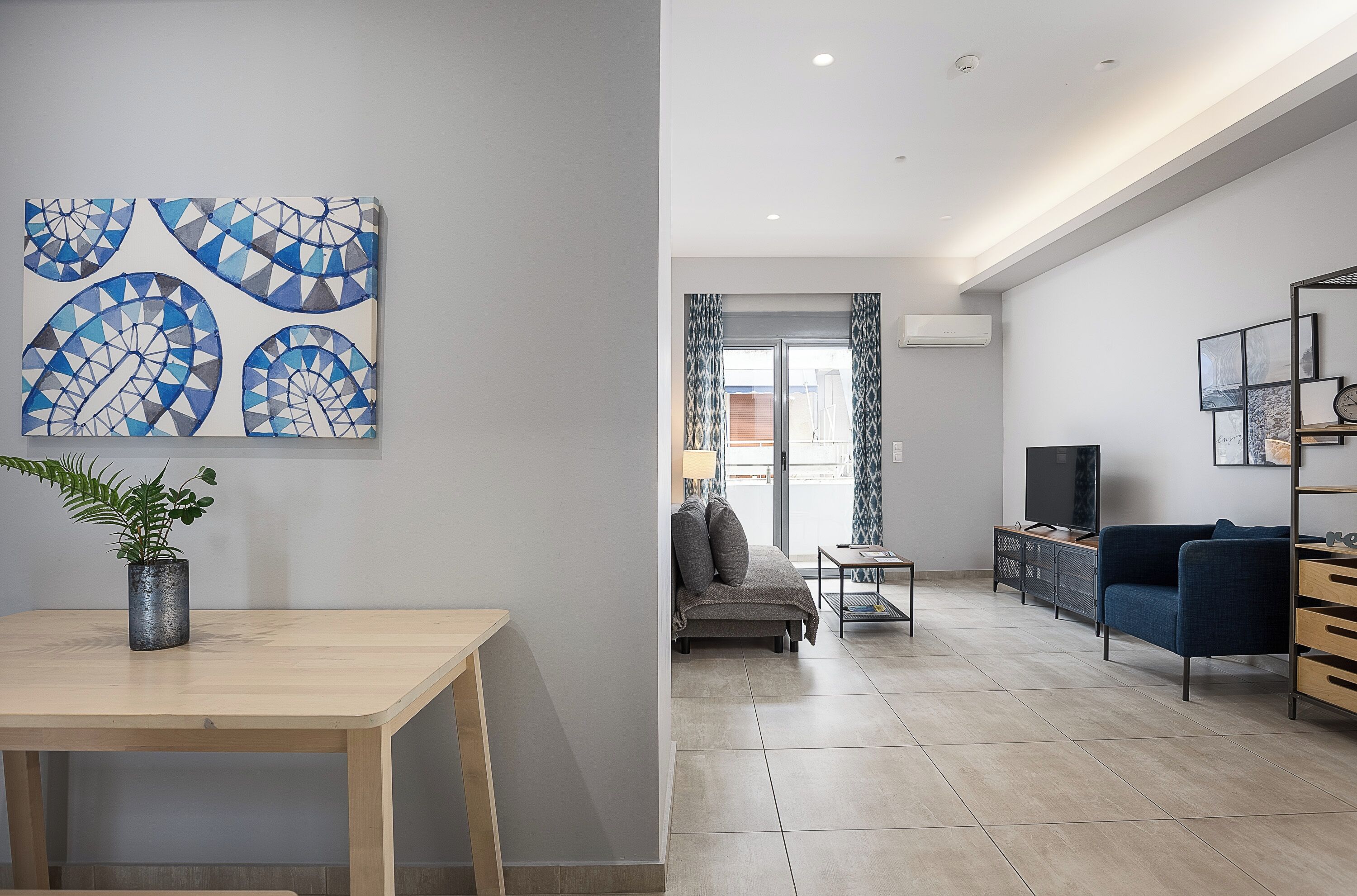 A modern living room with a wooden table, a blue abstract painting, a sofa, a TV, and a plant.