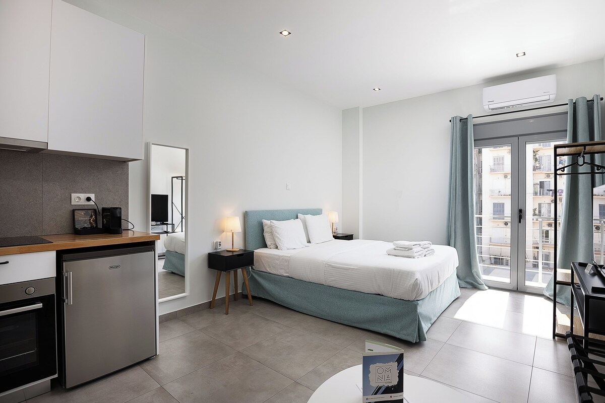 A modern hotel room with a kitchenette, a bed with a blue headboard, a bedside table with lamps, and a view through the glass door.