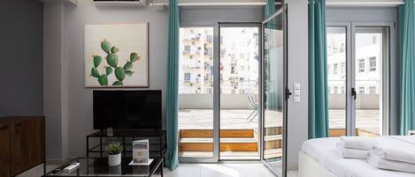A modern living room with a flat-screen TV, a glass coffee table, a sofa, and a bed. There is a sliding glass door leading to a balcony with a view of buildings.