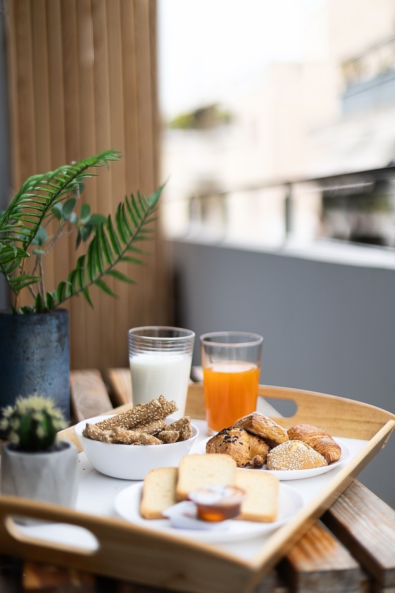 A tray with breakfast items including bread, croissants, and a glass of orange juice, with a plant and a glass of milk in the background.