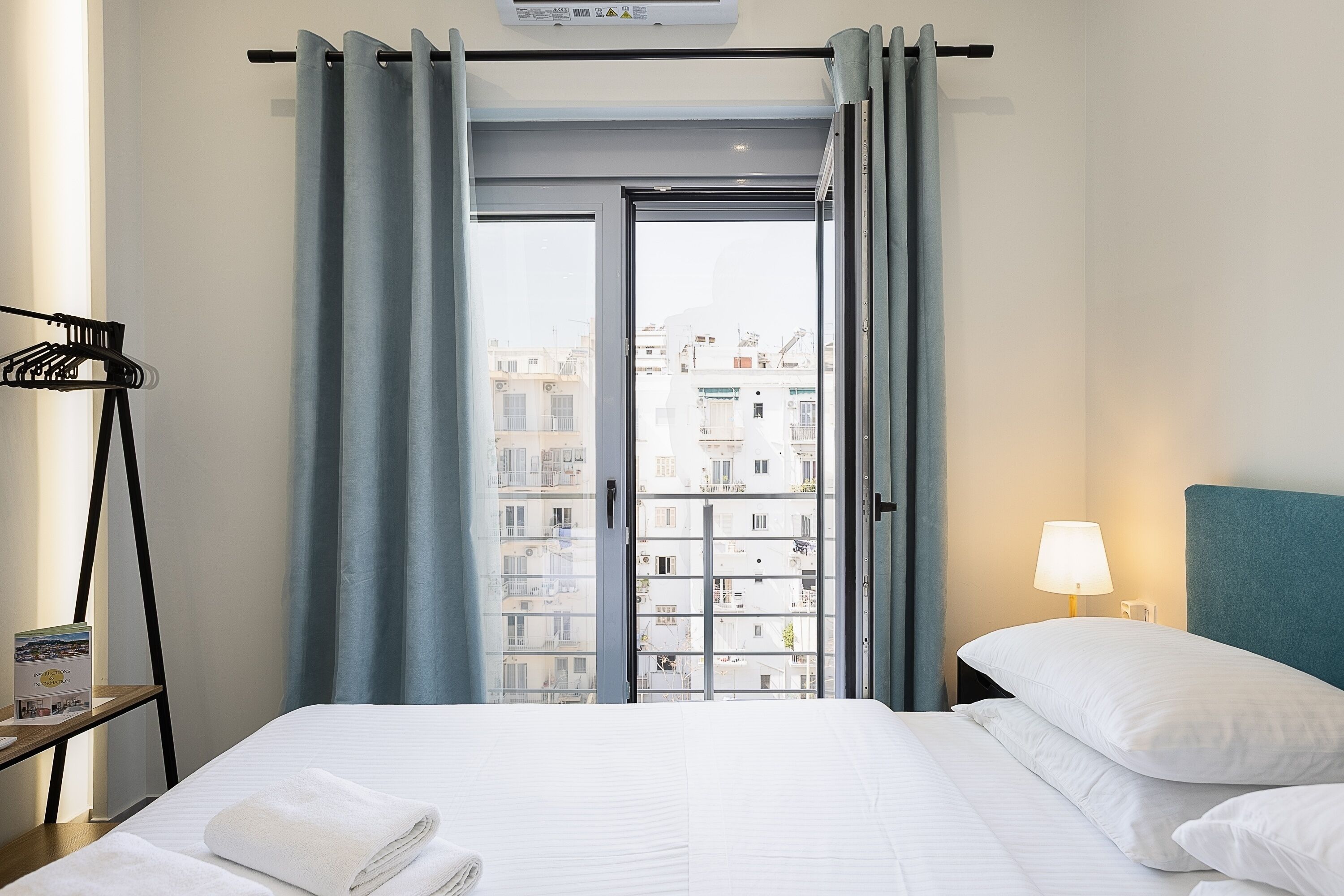 Ein Hotelzimmer mit einem Bett, einem Nachttisch mit Lampe, einem Fenster mit Vorhängen und Blick auf Gebäude.