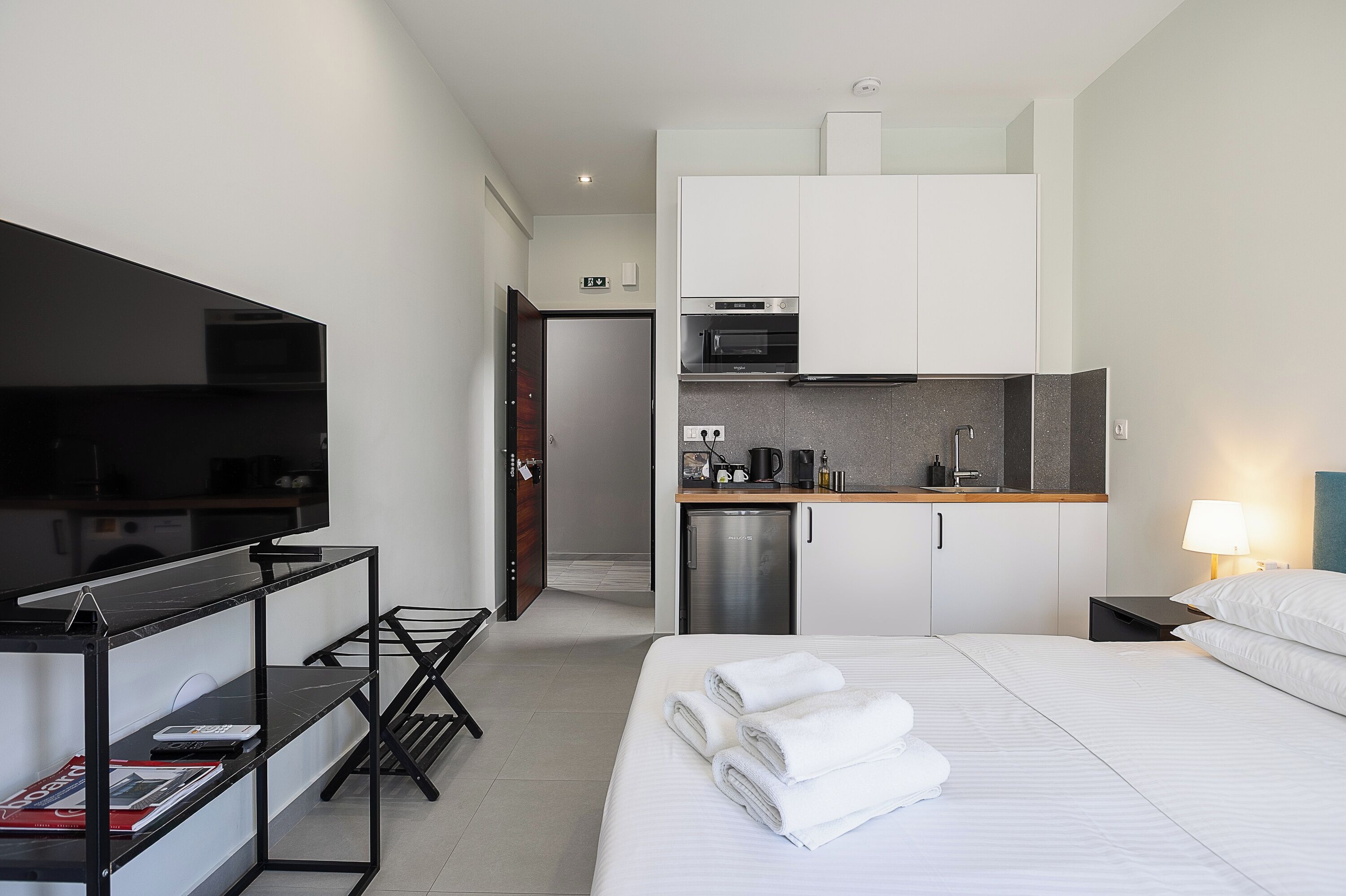 A hotel room with a bed, a TV, a desk with a chair, a kitchenette with a microwave, and a door leading to another room.