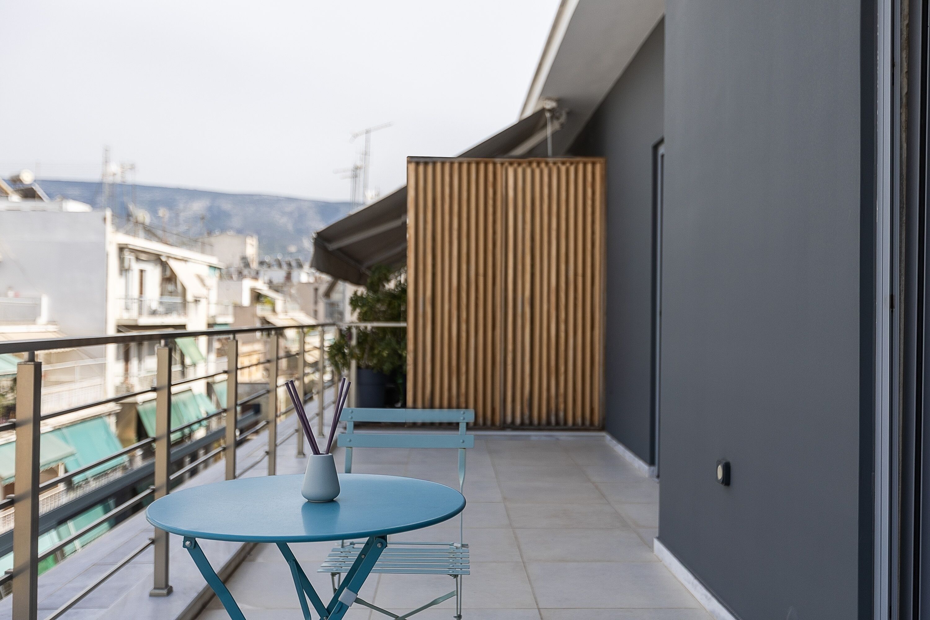 Un balcon avec une table et des chaises bleues, une cloison en bois et une vue sur des immeubles et l’eau.
