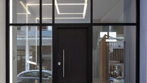 A modern building entrance with a glass door, a dark door, and a light fixture above. - The Omnia Collection Pagrati (Athens)