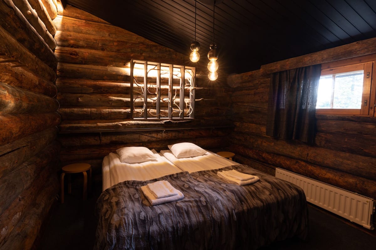 Una habitación tipo cabaña de madera con una cama, dos taburetes, un ventanal con cortinas y una lámpara fijada a la pared.