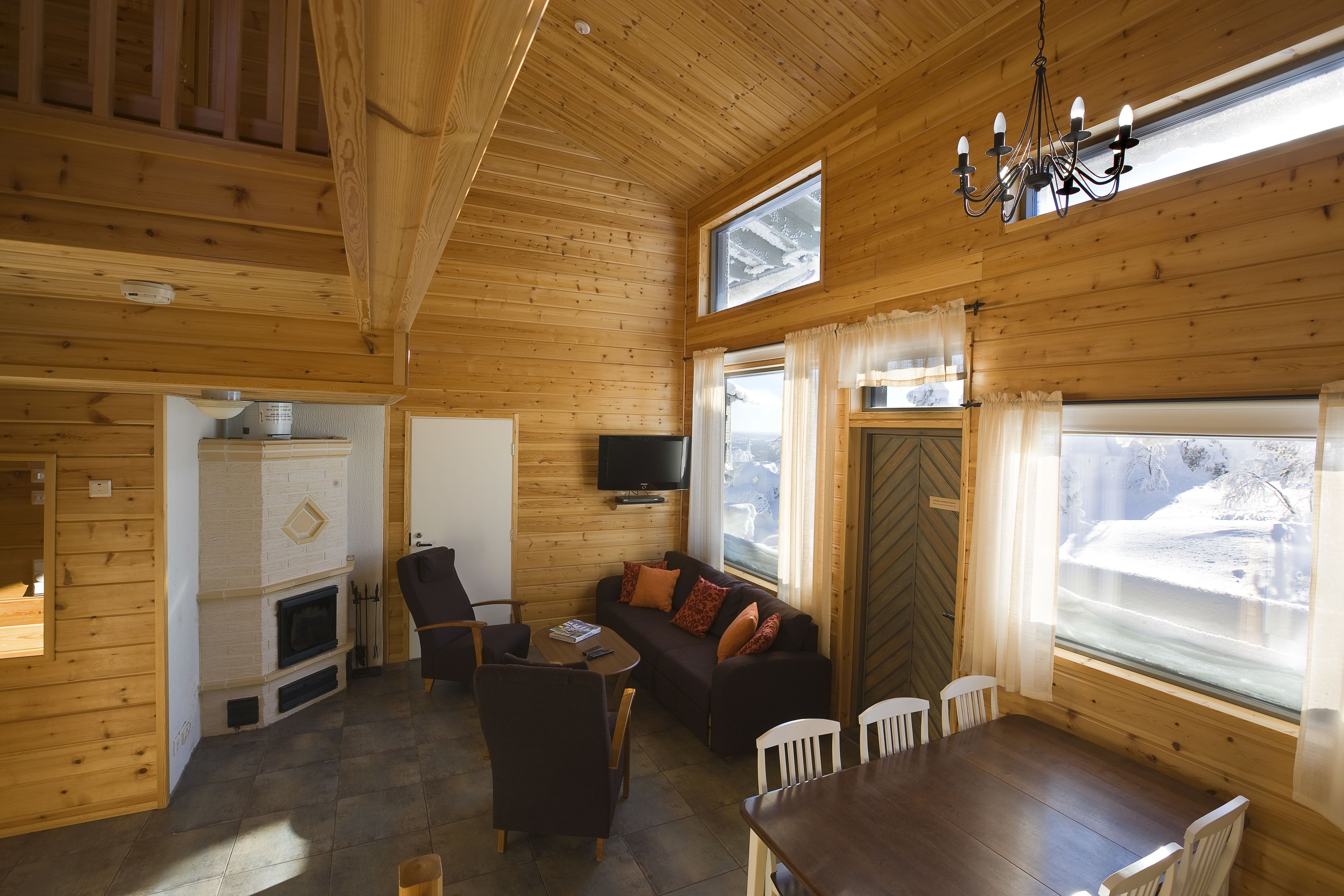 A wooden cabin interior with a fireplace, sofa, dining table, and large windows.