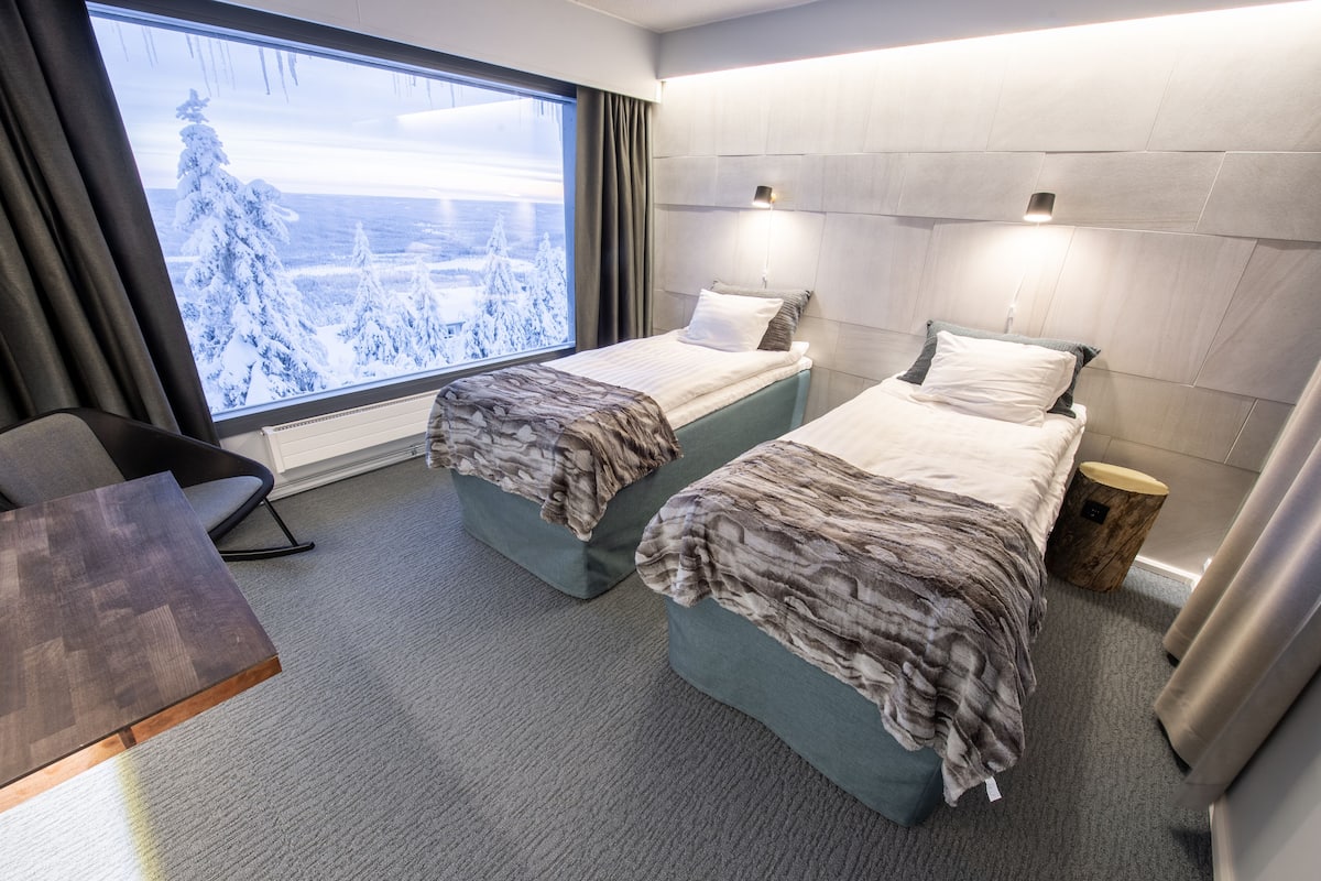 Habitación de hotel con dos camas, un escritorio y un ventanal con vistas nevadas.