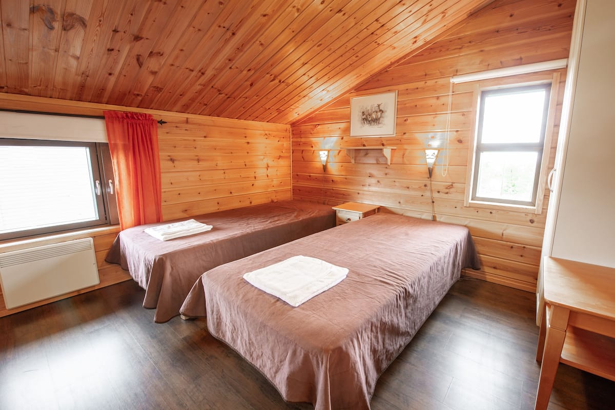 Una cabaña con dos camas, una ventana con cortinas, un radiador y un cuadro en la pared.
