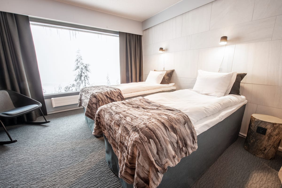 Habitación de hotel con dos camas, una silla, ventana con cortinas y vista a árboles.