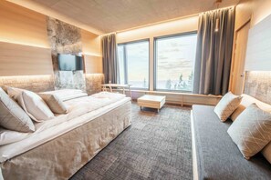 A modern hotel room with a large bed, a sofa, a TV mounted on a stone wall, and a view of the outdoors through large windows. - Hotel Iso-Syöte (Pudasjärvi)