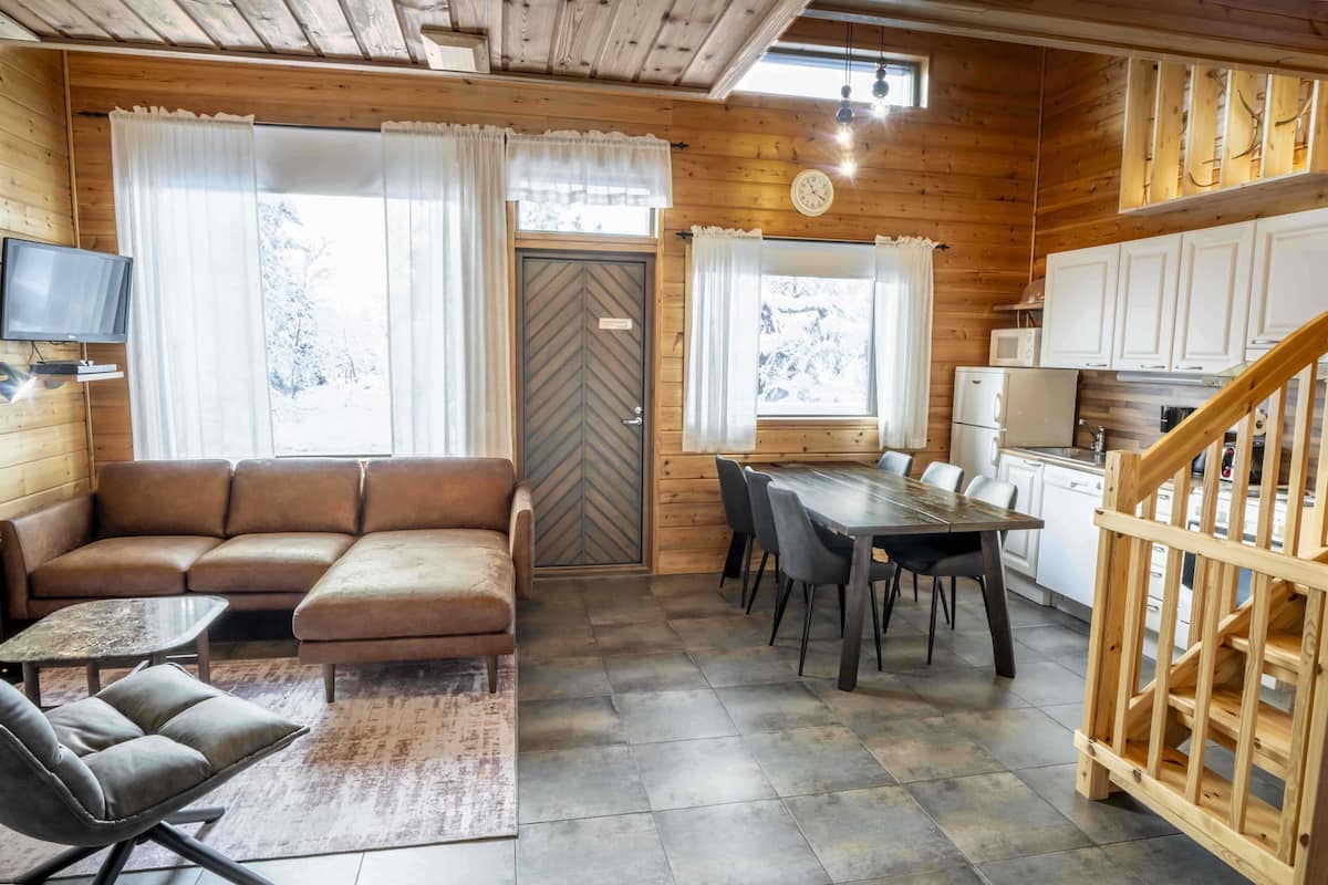 Un interior acogedor de cabaña con detalles de madera, un sofá, una mesa de comedor con sillas, una zona de cocina y un televisor montado en la pared.