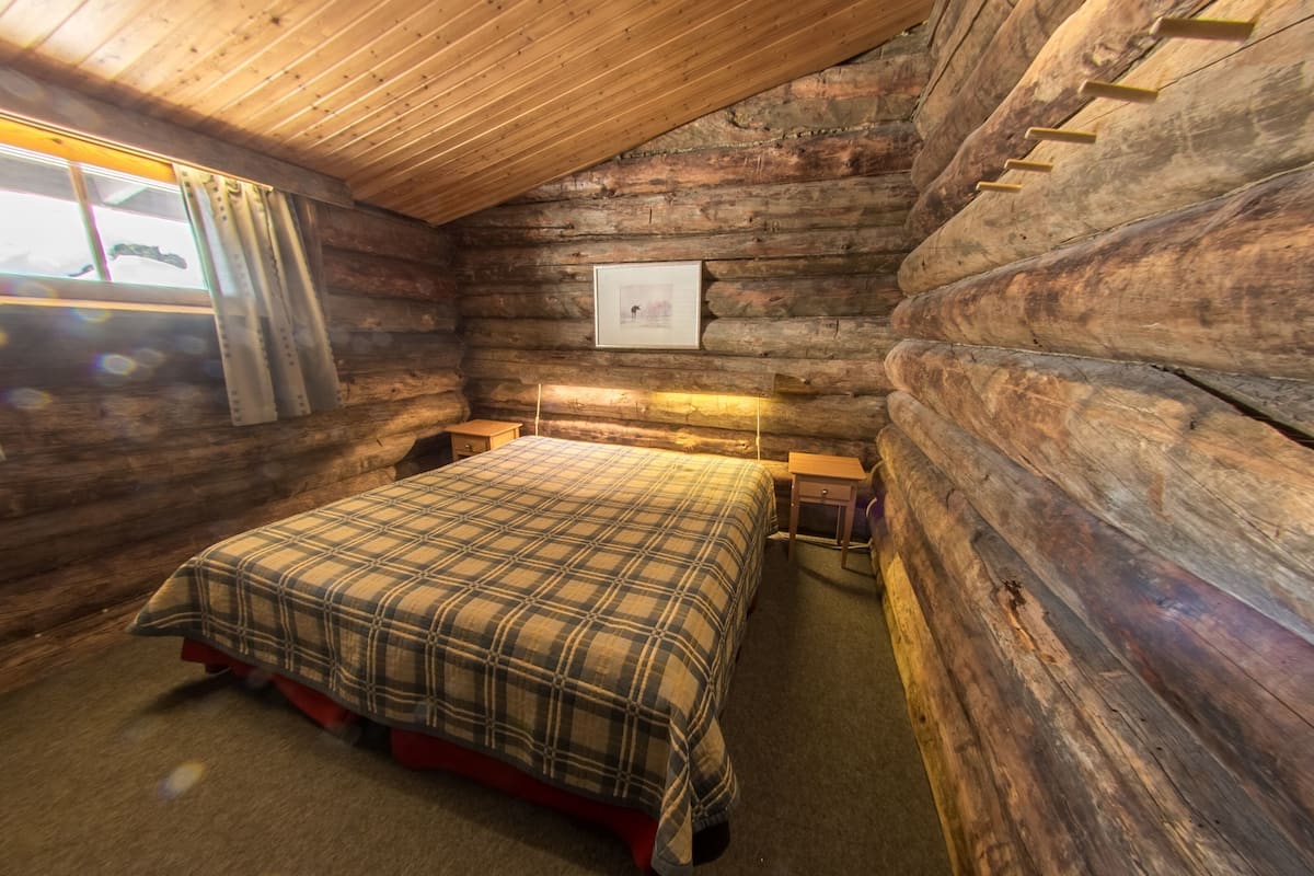 Un dormitorio tipo cabaña de troncos con una cama, una mesita de noche, un cuadro enmarcado en la pared y una ventana con cortinas.