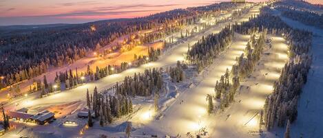 A snow-covered ski resort with illuminated ski runs, a parking area, and a lodge.