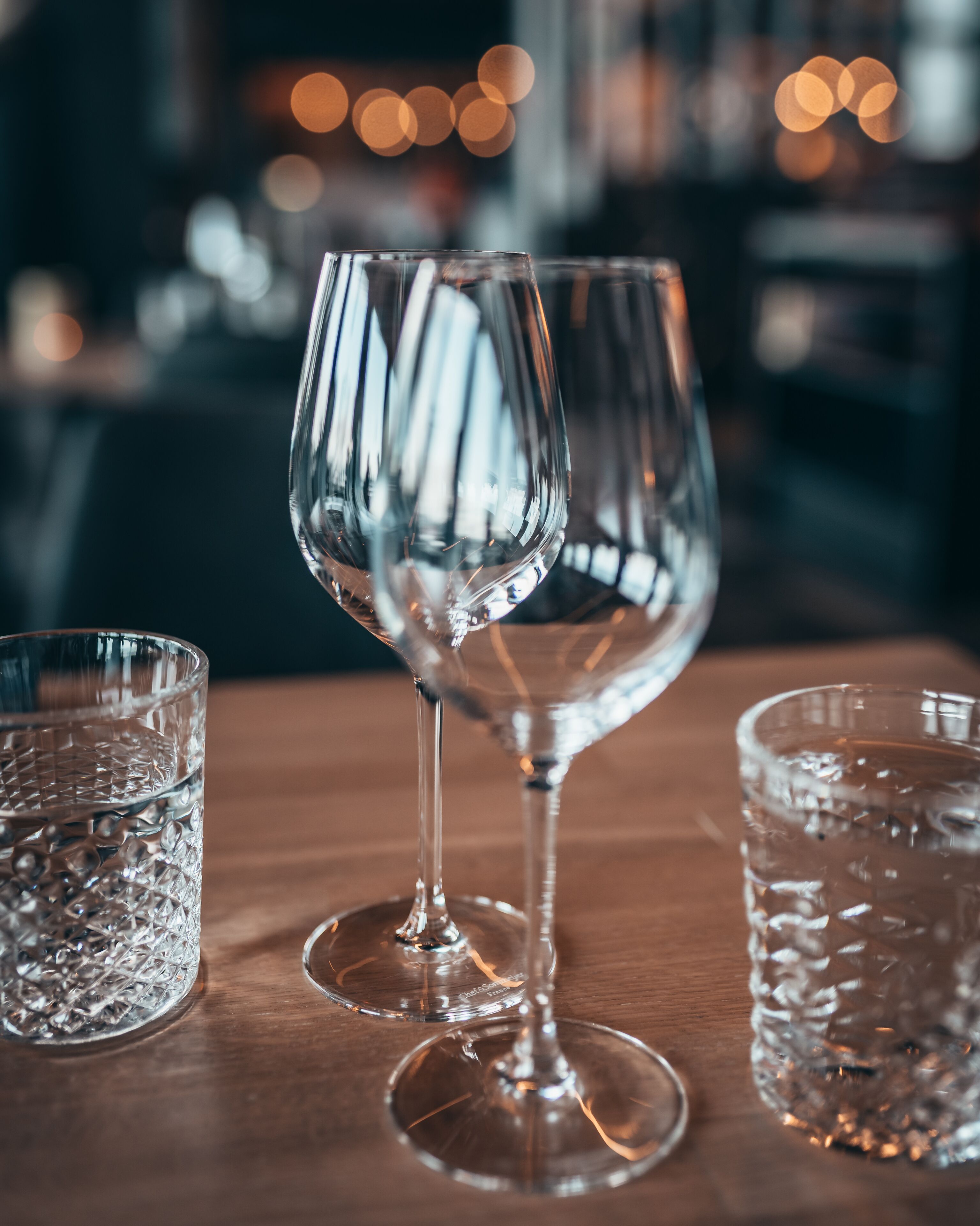 Trois verres à vin en verre transparent, posés sur une table en bois, avec un arrière-plan flou de lumières.