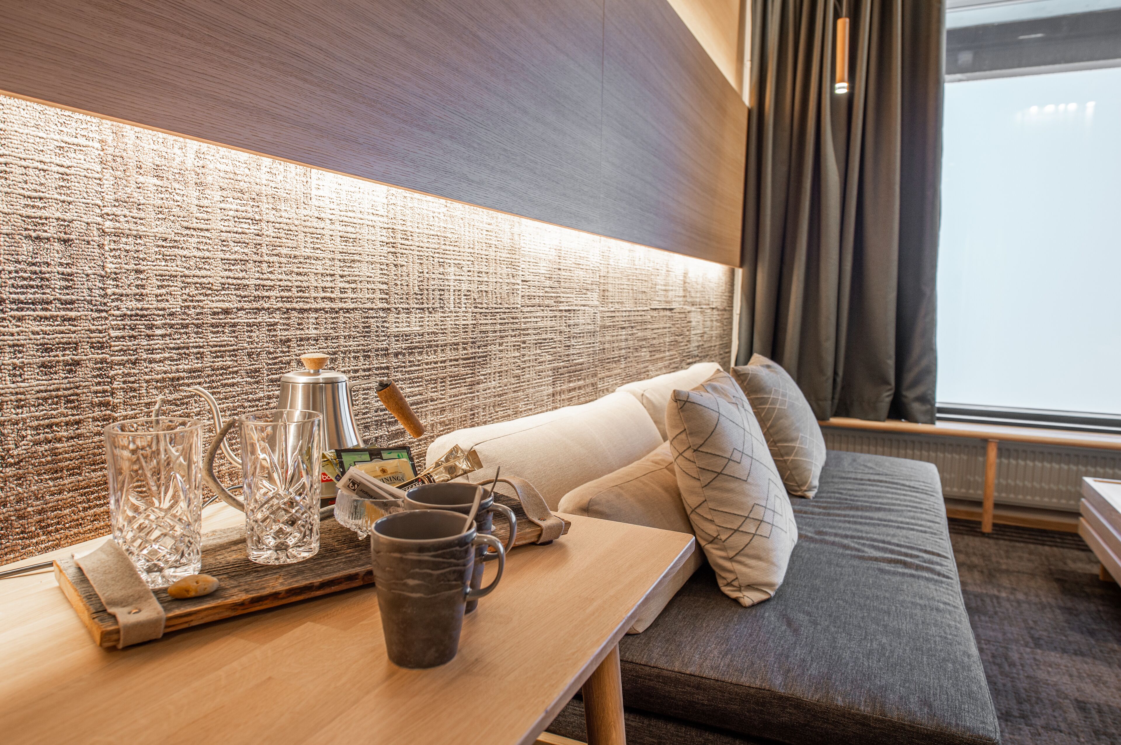 A modern room with a textured wall, a sofa with cushions, a table with a tray holding glasses and a mug, and a window with curtains.