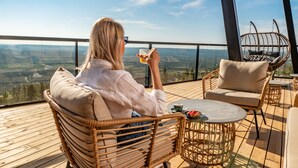 Une personne est assise sur une chaise en osier, sur une terrasse en bois, près d’une table et d’une chaise suspendue, et elle a une vue imprenable sur un paysage magnifique.