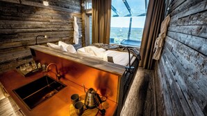 Une chambre de style cabane avec un intérieur en bois, un lit, un lavabo avec une bouilloire et une fenêtre donnant sur un paysage.