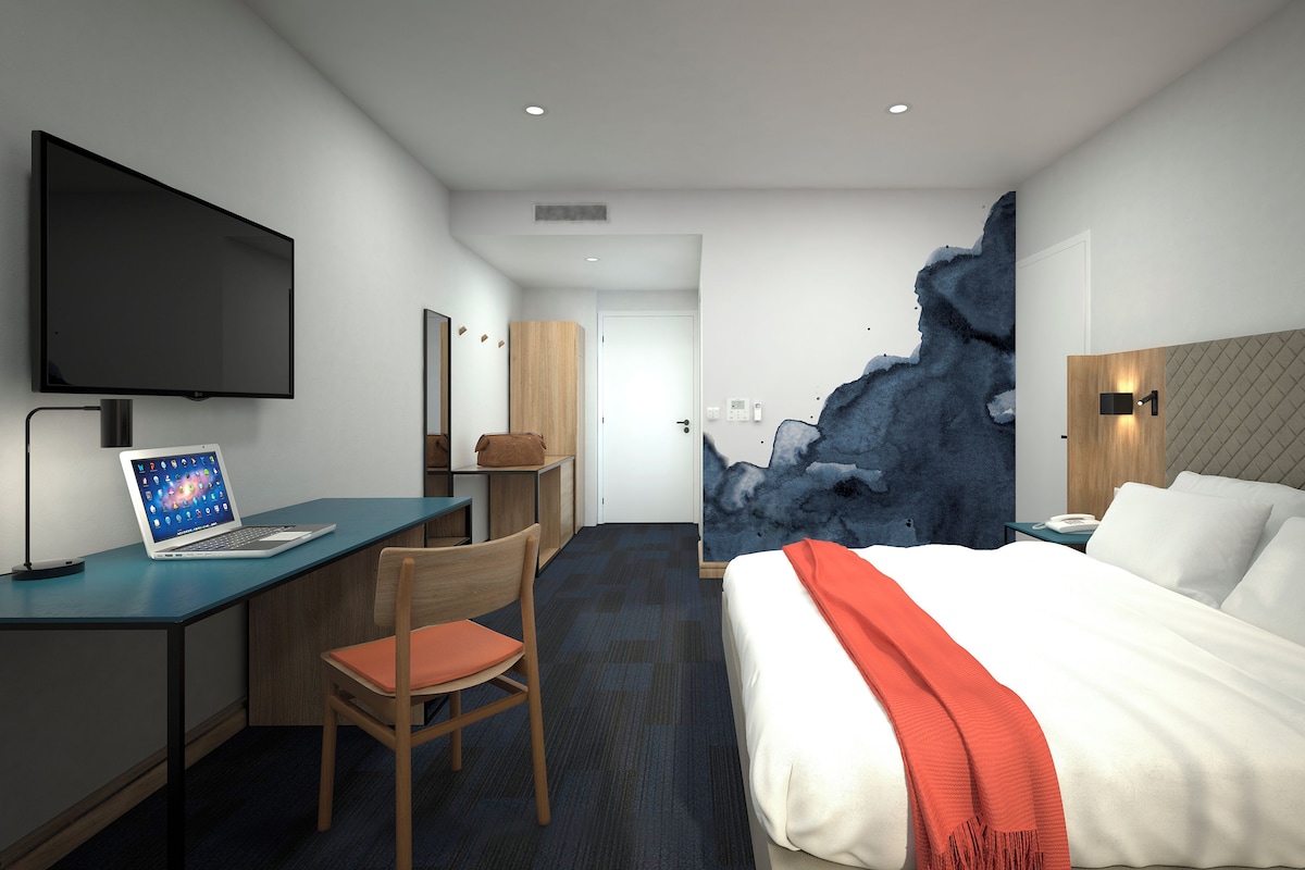 A hotel room with a bed, a desk with a laptop, a chair, a TV, and a unique wall feature.