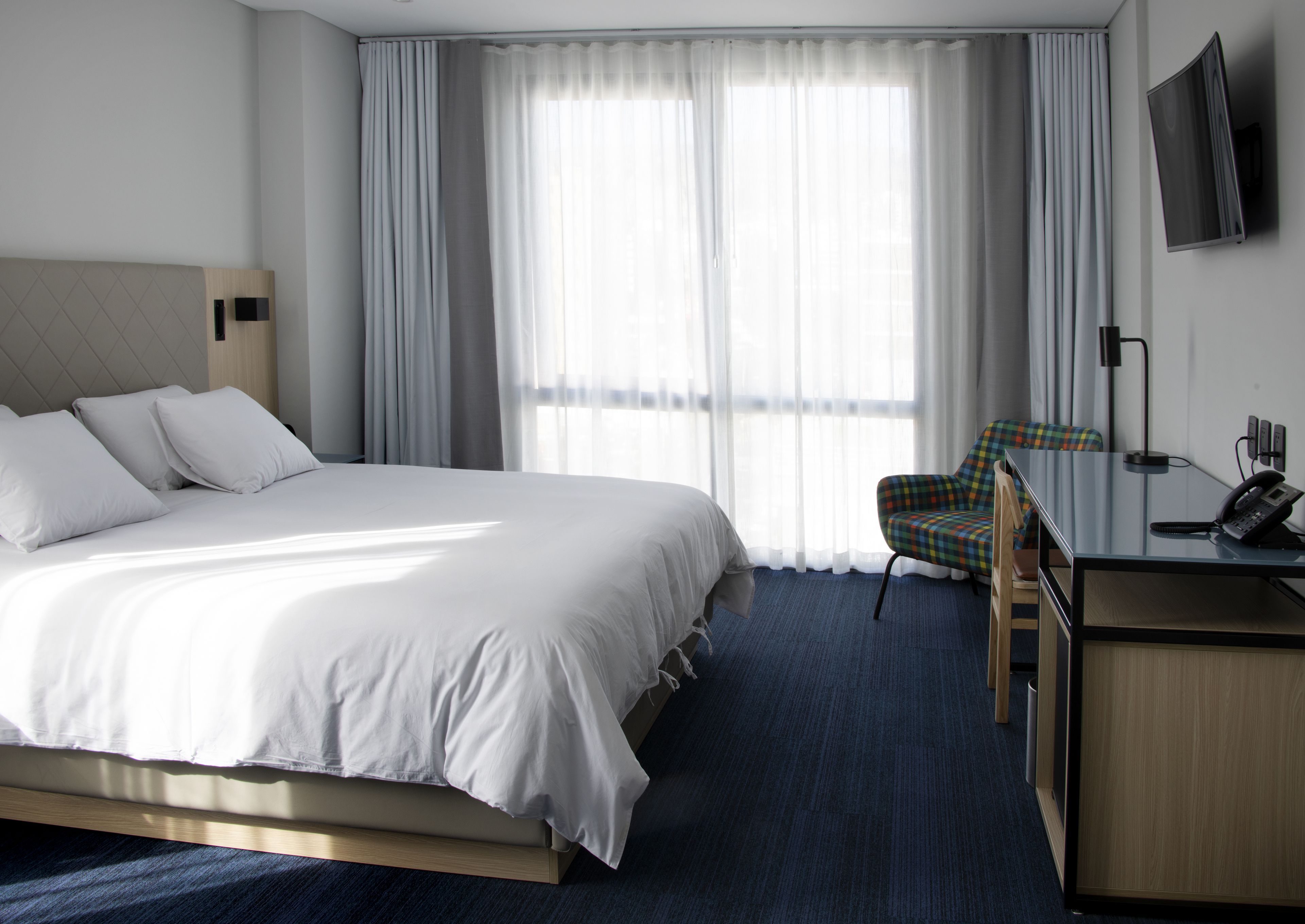 A hotel room with a large bed, a desk with a telephone, a chair, and a TV mounted on the wall.