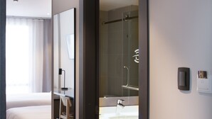 A hotel room with a bed, a desk, a bathroom with a sink and mirror, and a shower.