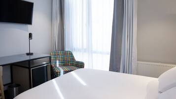 A hotel room with a large bed, a desk with a flat-screen TV, a chair, and a window with sheer curtains.