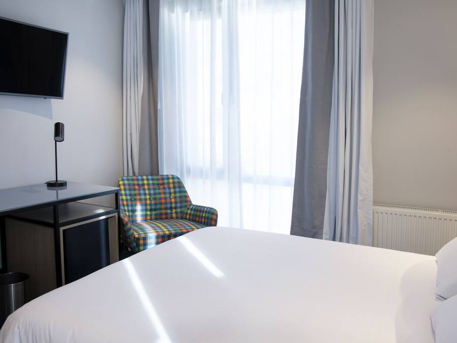A hotel room with a large bed, a desk with a flat-screen TV, a chair, and a window with sheer curtains.