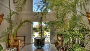 Un espace intérieur vaste et lumineux, agrémenté de grands pots de plantes, d’un banc en bois et offrant une vue sur l’extérieur à travers une porte vitrée.