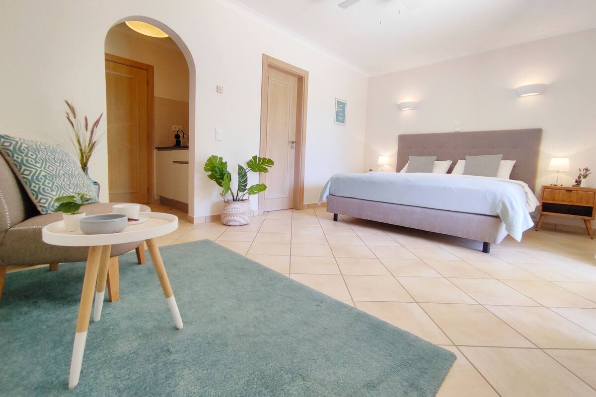 Une chambre à coucher comprenant un lit, des tables de chevet, une petite table avec un bol et une tasse, une plante en pot et une porte donnant sur une autre pièce.
