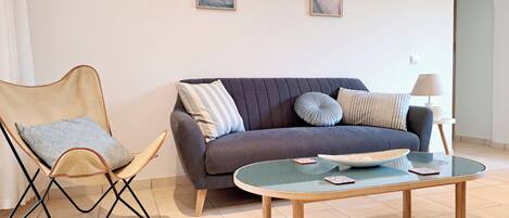 A modern living room with a grey sofa, a wooden chair, and a glass coffee table. There are three framed pictures on the wall.
