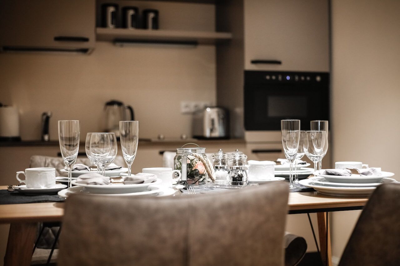 A dining table set with wine glasses, plates, and cups, with a kitchen in the background.