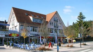 A building with a red-tiled roof, multiple windows, and a storefront with a sign that reads "RESTAURANT." There are outdoor tables and chairs in front of the building. - Appartementhaus Am Roten Platz (Titisee-Neustadt)