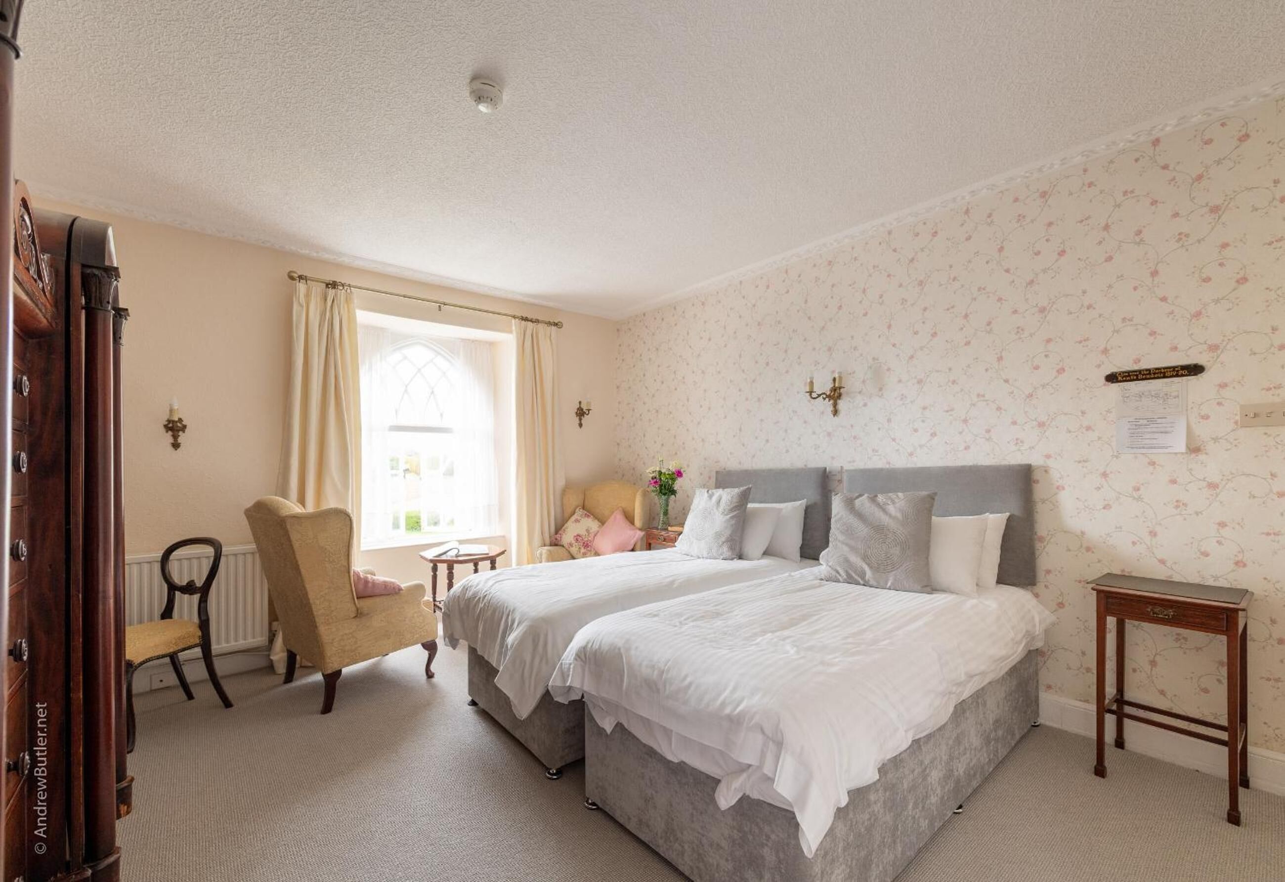 A hotel room with two beds, a chair, a small table, and a window with curtains.