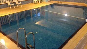 An indoor swimming pool with clear blue water, a tiled deck area, and a set of stairs leading into the pool.