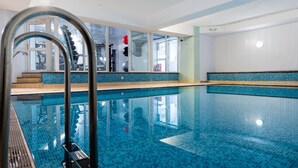 Una piscina techada con paredes de vidrio transparente, piso de baldosas y escalera metálica.