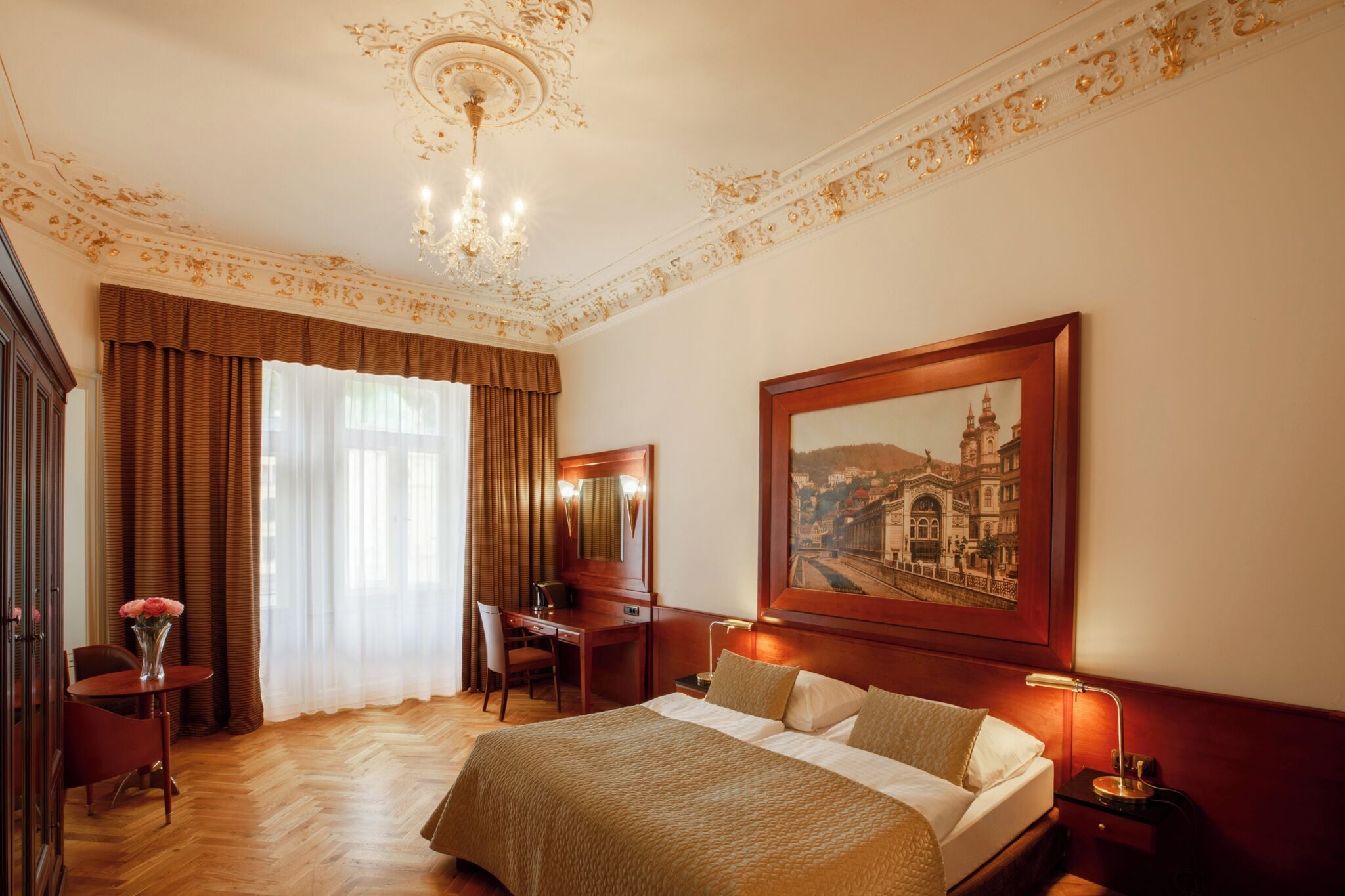 A hotel room with a large bed, a desk, a chair, a desk lamp, a nightstand, a vase with flowers, and a painting on the wall.