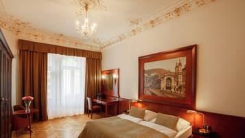A hotel room with a large bed, a desk, a chair, a desk lamp, a nightstand, a vase with flowers, and a painting on the wall.