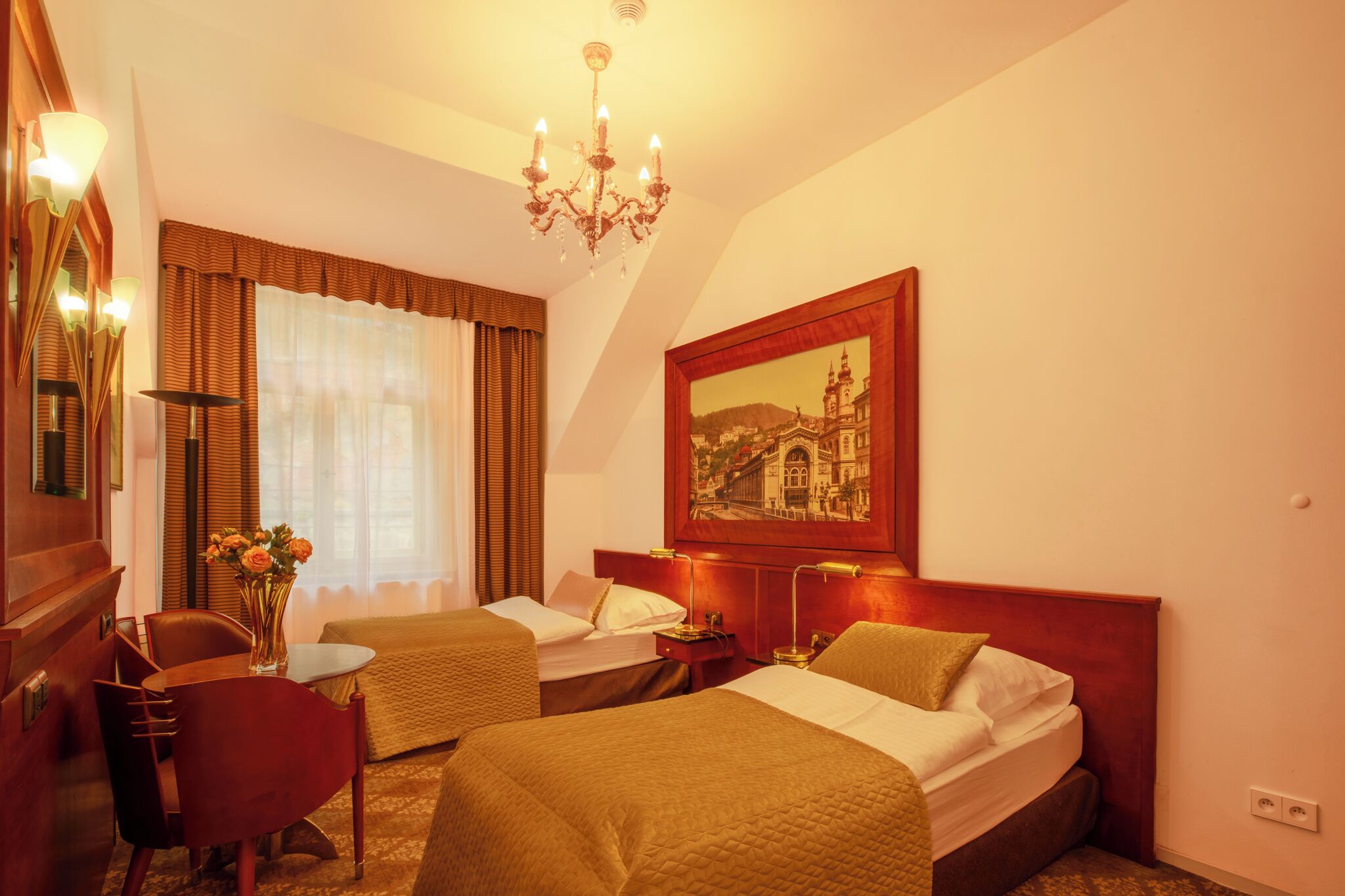 A hotel room with two beds, a desk, a chair, a chandelier, and a painting on the wall.