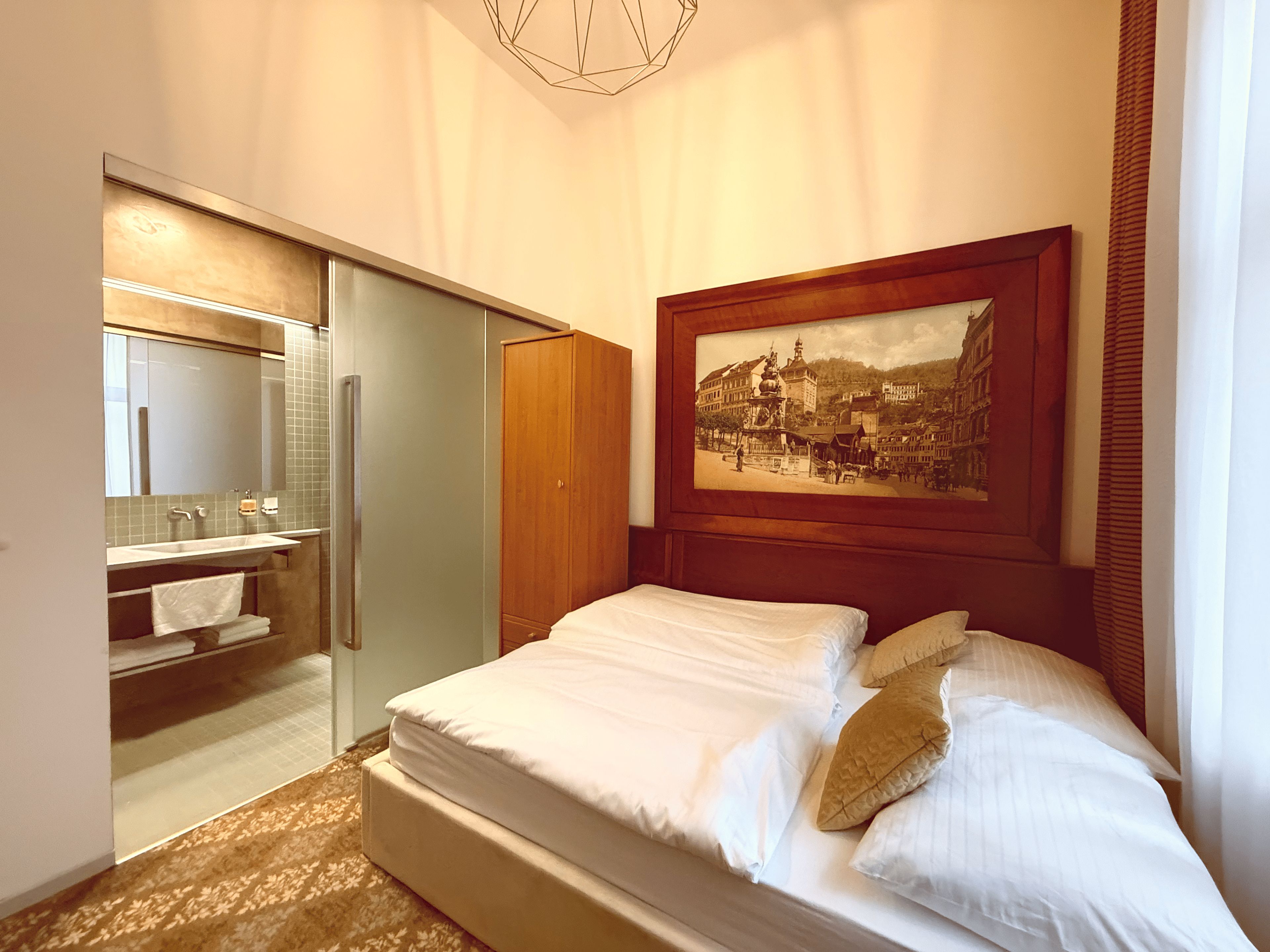 A hotel room with a bed, a painting, a bathroom with a mirror, and a wardrobe.