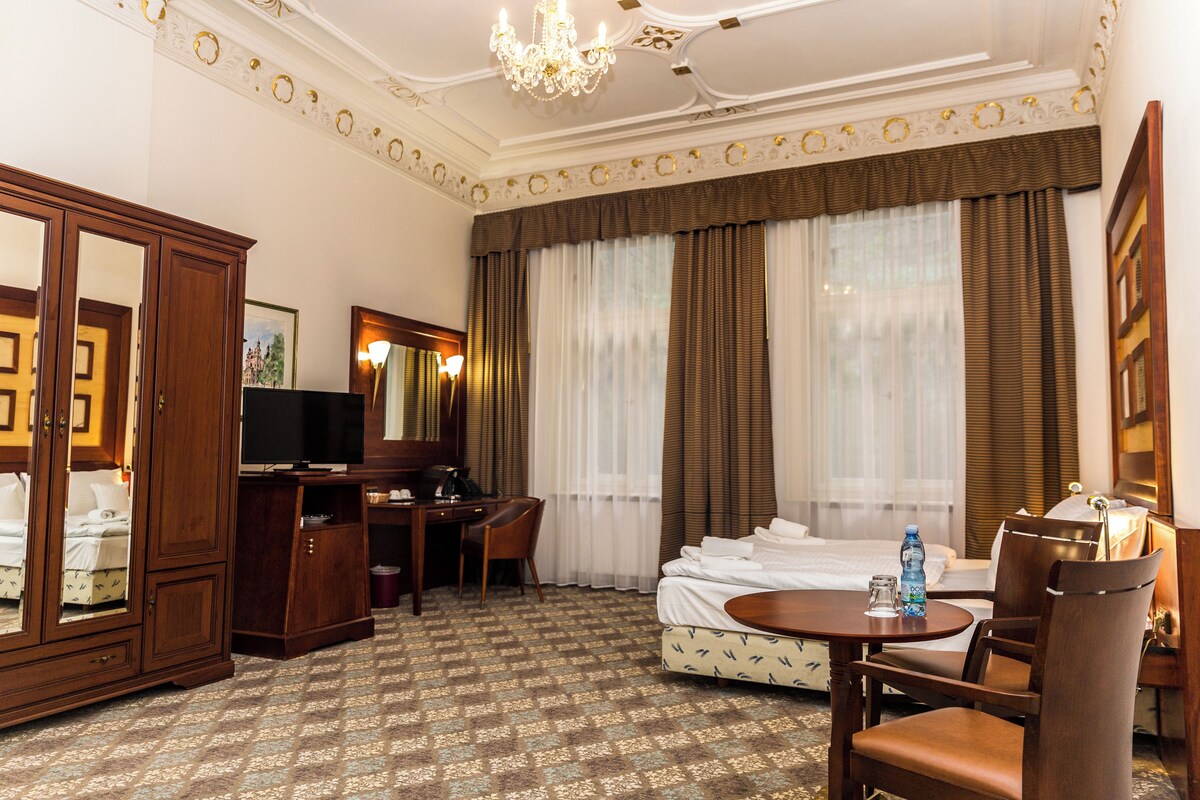 A hotel room with a bed, a desk with a chair, a wardrobe, a television, and a chandelier.