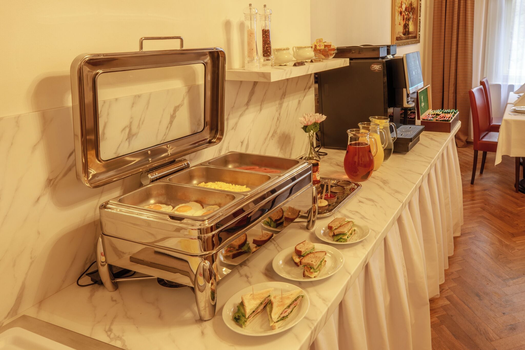A buffet setup with a variety of food items and beverages, a refrigerator, and a computer on a counter.